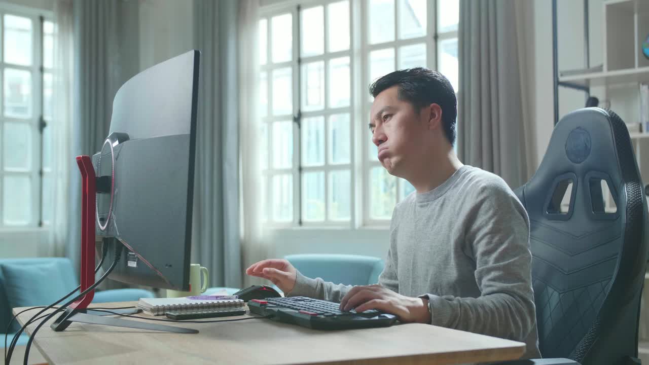 Asian Man In Long Sleeved T-Shirt Being Tired While Using The Desktop Computer For Working At Home.