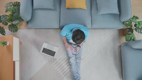 top view zoom out shot of a man using smartphone while working on a laptop on the floor in front of a sofa at home