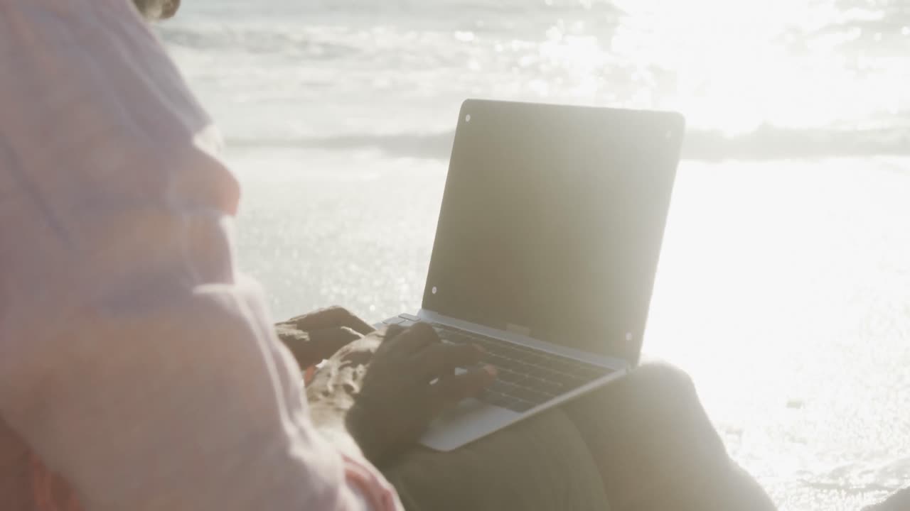 시니어 아프리카계 미국인 남자가  ⁇ 니 비치에서 노트북을 사용하고 앉아 있습니다.