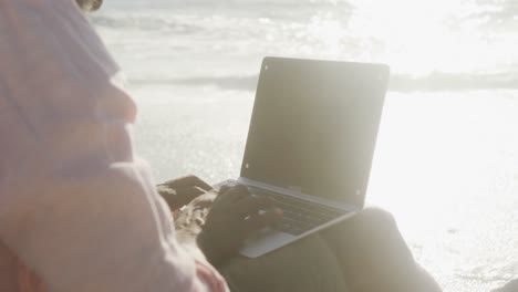 hombre afroamericano sénior sentado y usando una computadora portátil en sunny beach