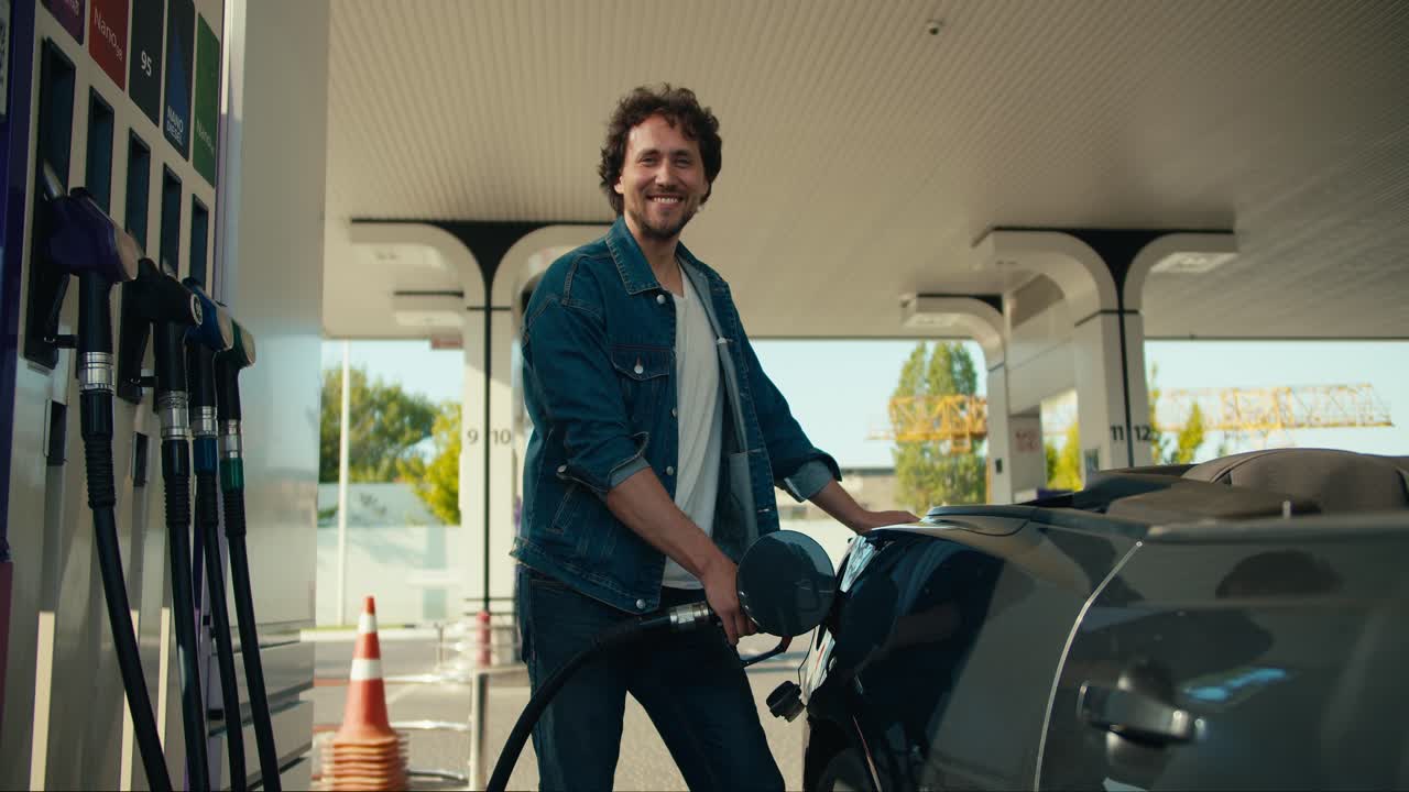 Portrait of a curly-haired brunette guy who refuels a gray convertible. Happy car ride, gas station stop