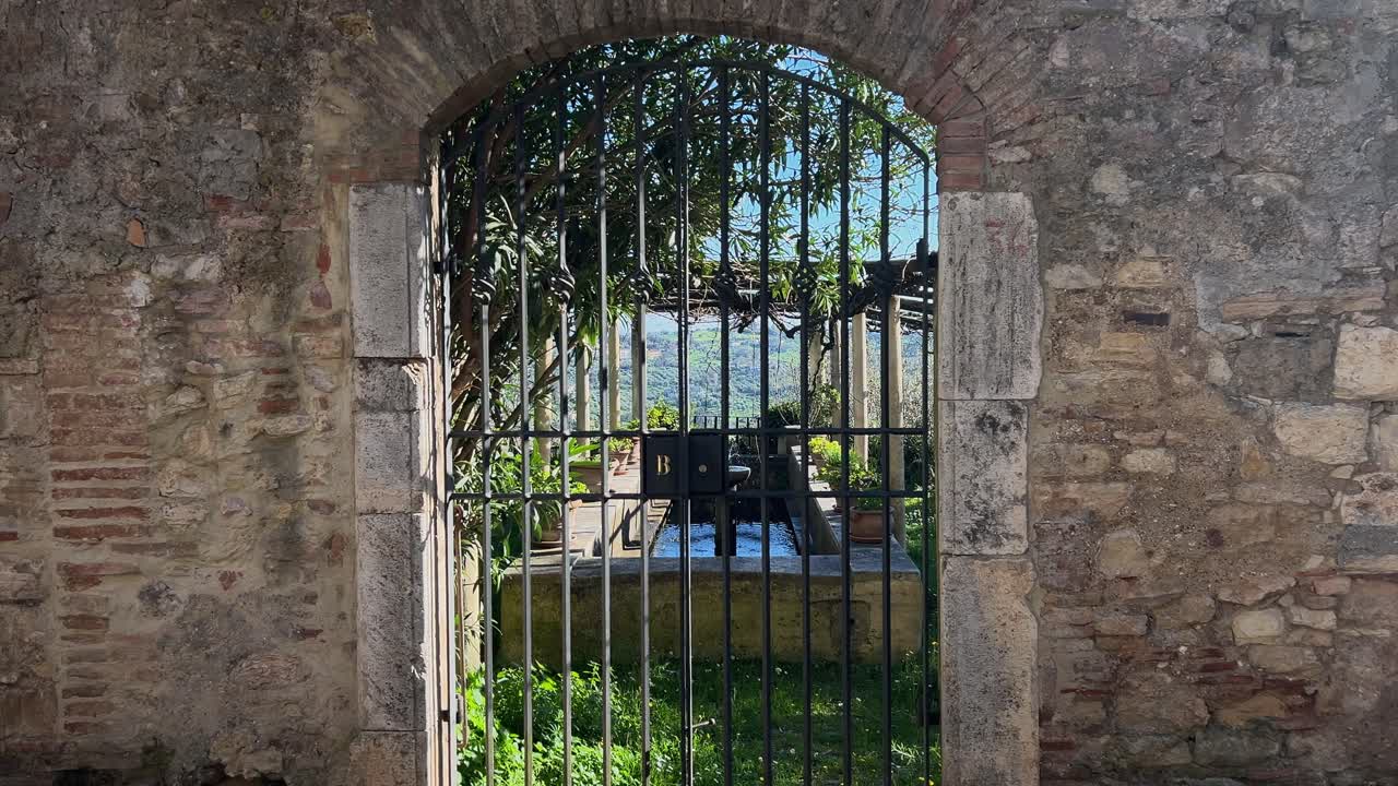 View through an old iron gate of a historic stone fountain under a pergola in a lush green garden in Amelia, Umbria, Italy. Secret garden concept