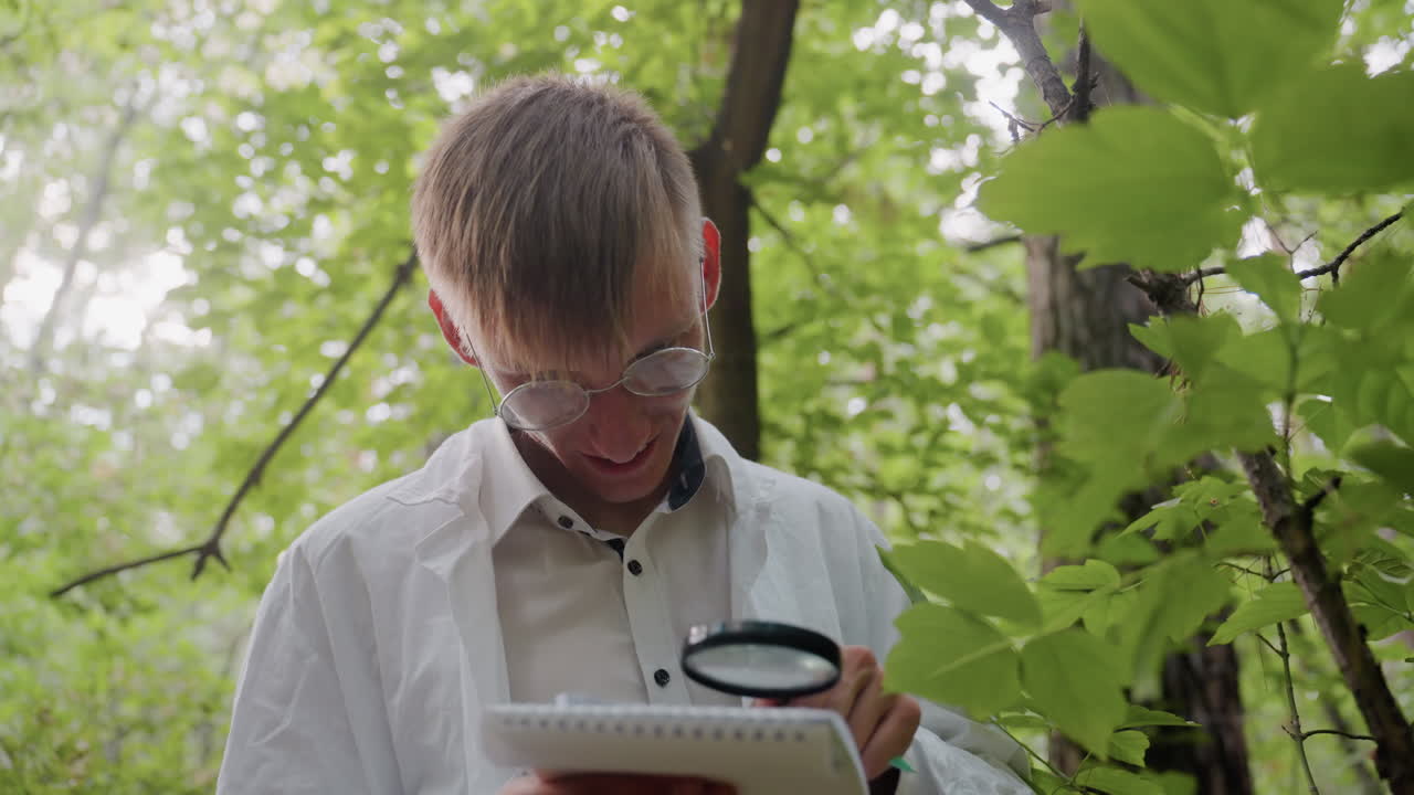 Young man in white coat and glasses blows breeze on microscope and observes carefully while conducting forest research, combining curiosity with scientific study surrounded by natural greenery
