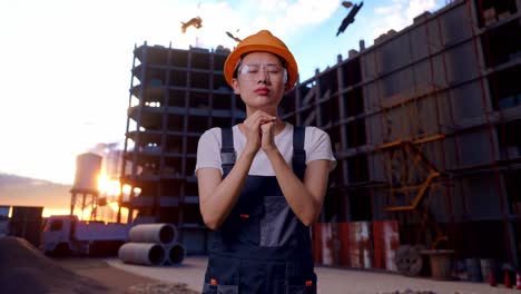 trabajadora asiática con gafas de protección y casco de seguridad orando por algo mientras está de pie en el sitio de construcción