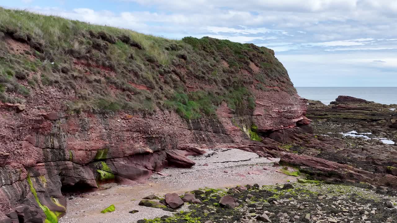 Camera slowly pans revealing red sandstone cliffs, rocky shore, seaweed, and overcast coastal landscape