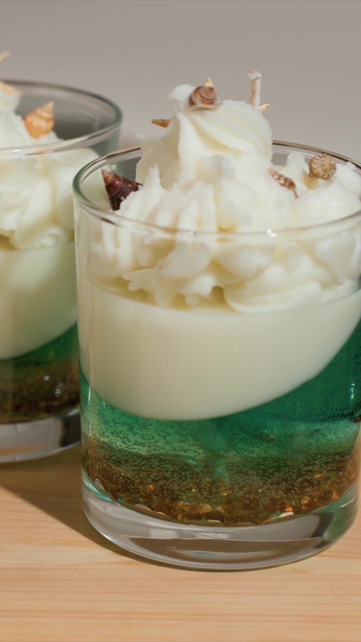 Close up of layered candles in glass with green and white wax topped with shells. Candles sit on wooden surface under soft lighting showcasing detailed textures like smooth cream colored top