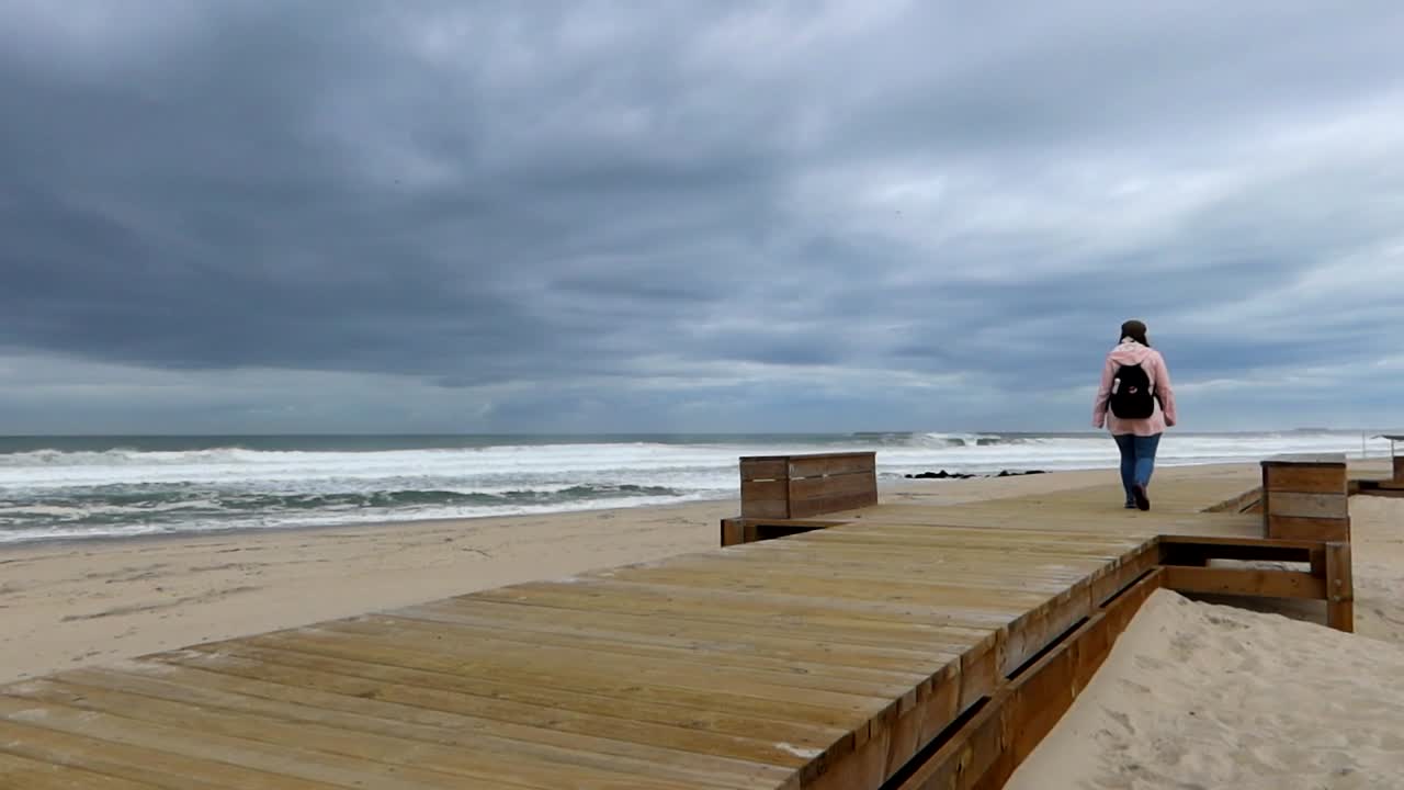 Solitary woman in raincoat strolls along Costa Nova's wooden path, Portugal