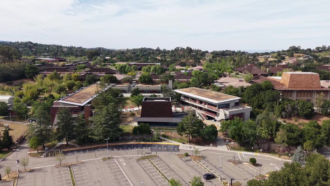 Vista descendente de dron capturando la arquitectura icónica y los tejados verdes de Foothill College en Los Altos Hills, California, mostrando el campus vacío y el exuberante paisaje circundante durante un día soleado.