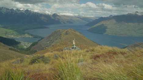 Woman-stepping-onto-rock-viewpoint-raising-arms-out-of-joy,-lake-Wakatipu