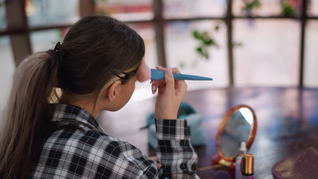 vista aérea de cerca de una dama sosteniendo un pincel de maquillaje, aplicando cuidadosamente el producto en la cara, mesa de madera con cosméticos, espejo que refleja la luz suave, creando un entorno de rutina de belleza elegante y enfocado