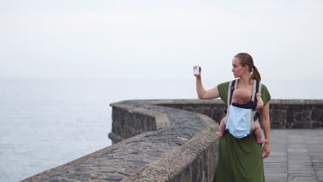 una joven madre con un bebé dispara en sus manos mirando el océano y dispara una impresionante vista de las olas en su teléfono inteligente