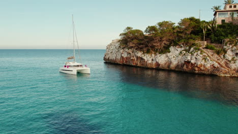 catamaran boat sailing in the blue sea with houses on the cliff in cala llombards, mallorca, spain