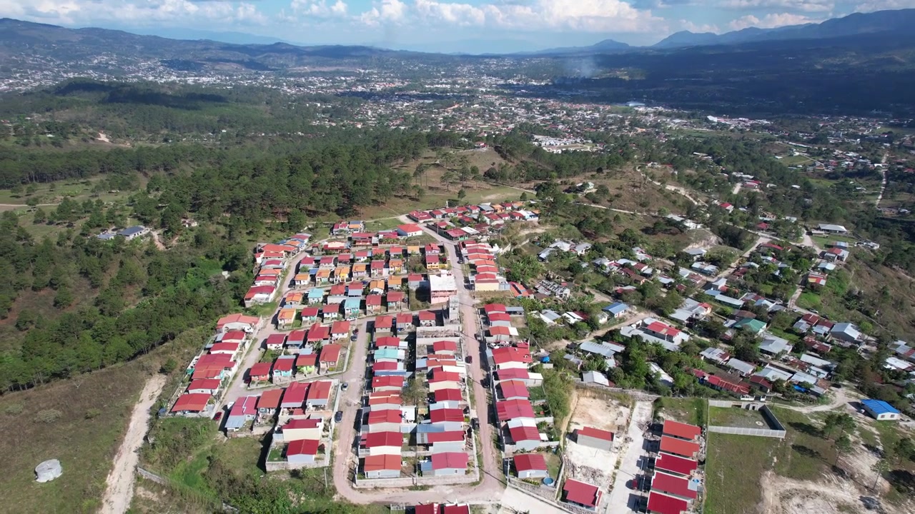 Una toma de dron a gran altitud revela un desarrollo residencial latinoamericano con aproximadamente 150 casas con techos de zinc rojos, ubicado entre un denso bosque verde y terreno abierto.