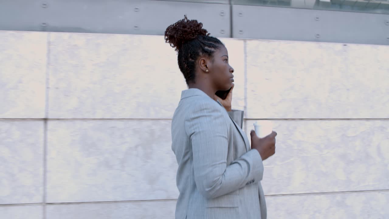 Dolly shot of a focused African-American businesswoman walking outside, holding coffee and using mobile phone