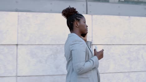 dolly shot of a focused african-american businesswoman walking outside, holding coffee and using mobile phone