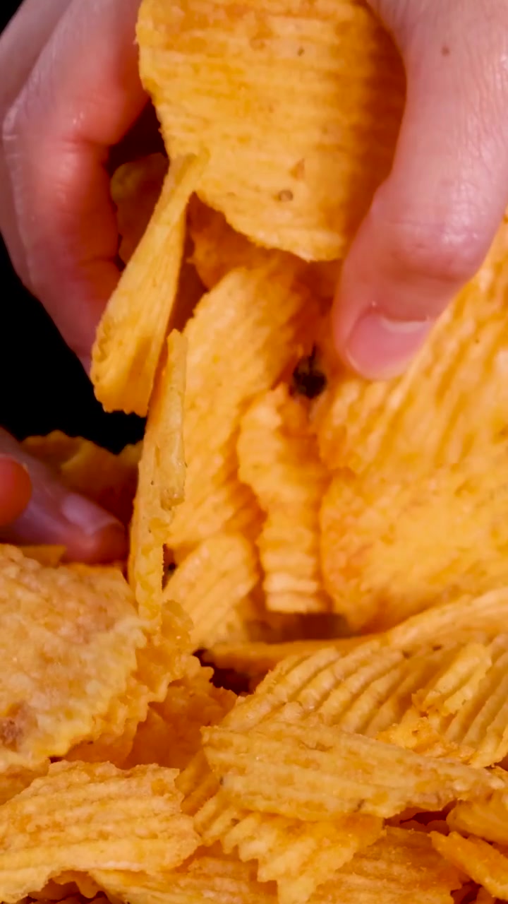 Uma mão alcança uma pilha de batatas fritas douradas e onduladas contra um fundo escuro.