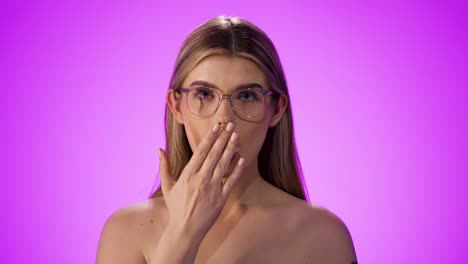 medium shot of a pretty young woman wearing glasses and holding her hand in front of her mouth with a startled look and looking at the camera in amazement or surprised look against a purple background