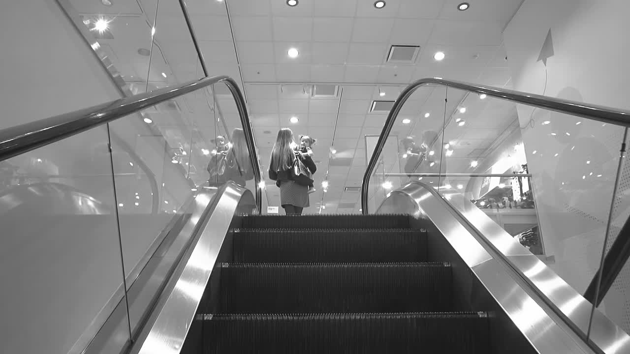 어린 아이를 가진 젊은 어머니는 단색 색의 느린 동작으로 옷 가게의 에스컬레이터에 올라갑니다.