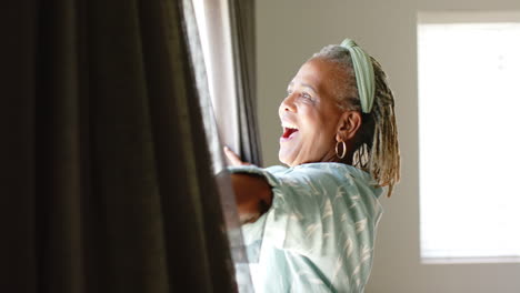 Mujer-Mayor-Afroamericana-Abriendo-Cortinas,-Mirando-Afuera