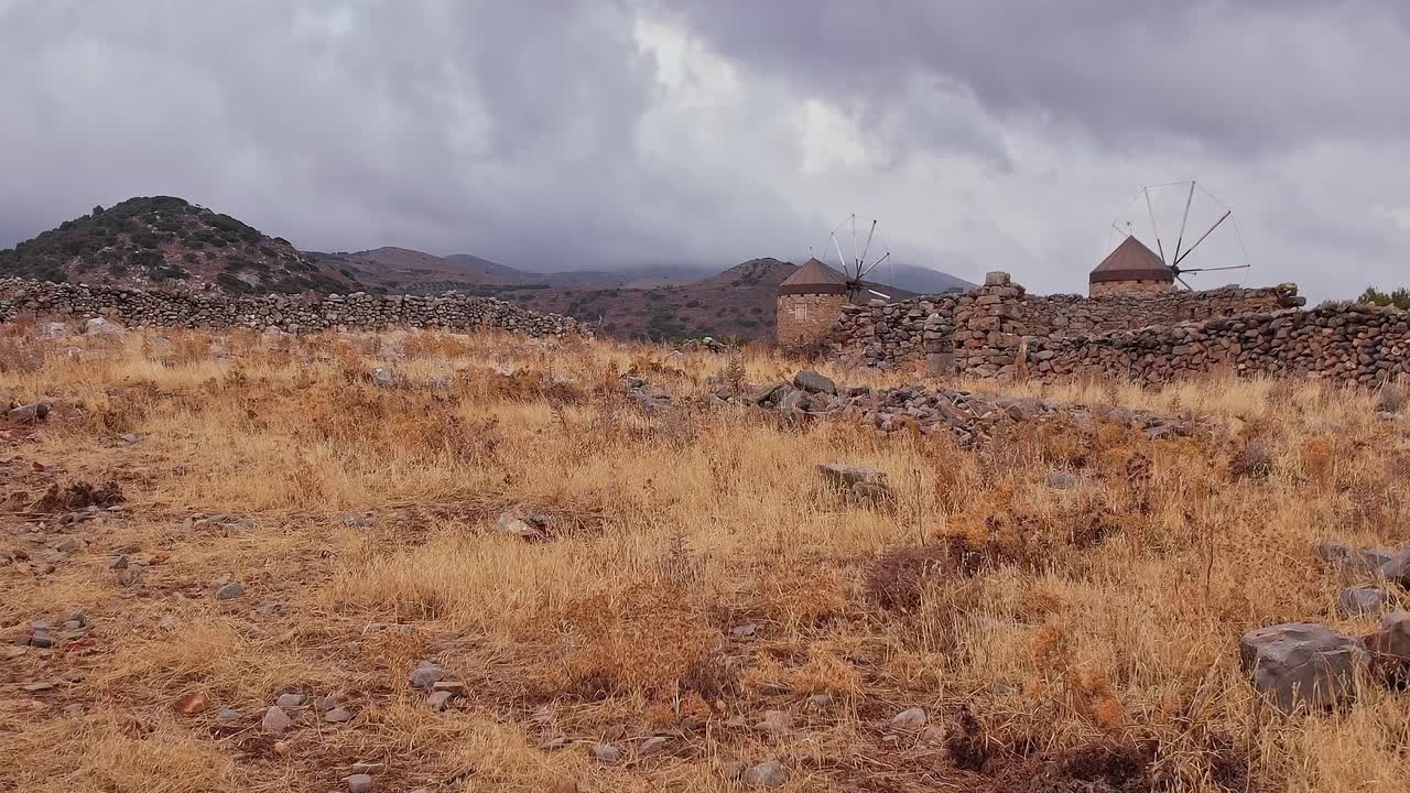 Vecchi mulini a vento e muri in pietra in un paesaggio montano arido sotto un cielo nuvoloso
