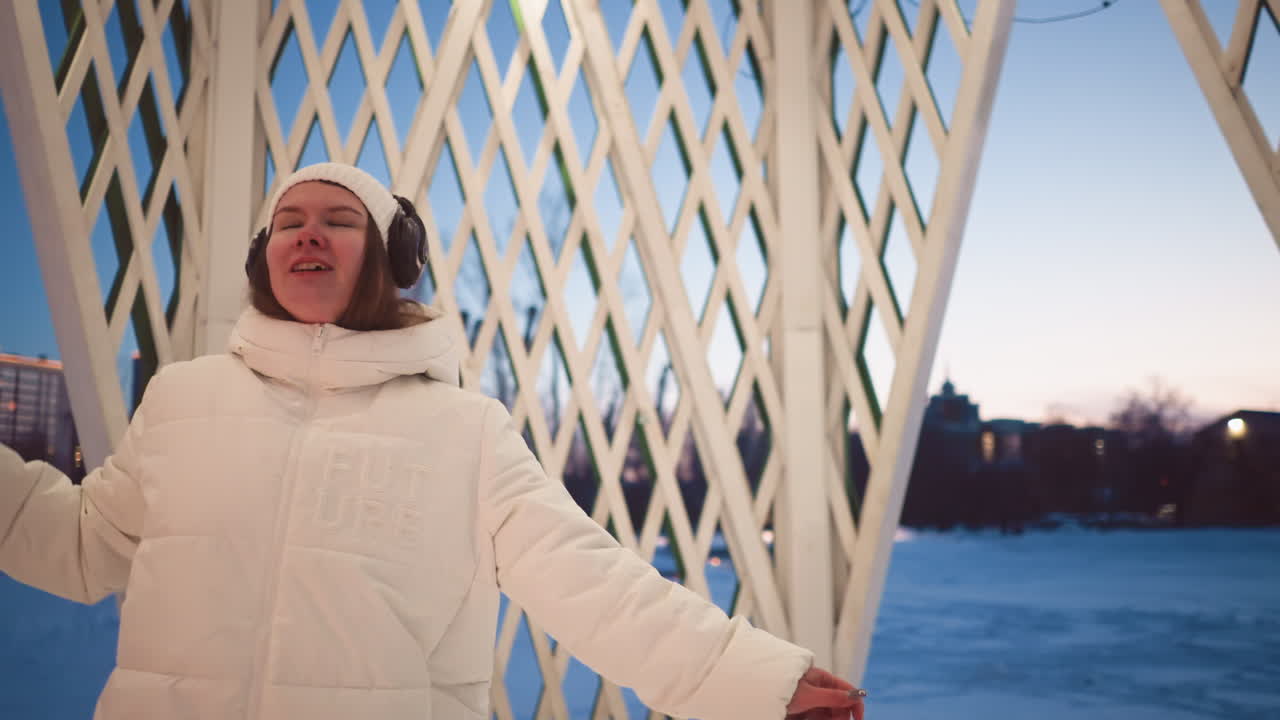 Close up of young girl spinning with eyes closed wearing headset in well lit snowy pavilion at dusk, arms raised in serene rhythm beneath bare trees, cool evening air enhancing joyful winter dance