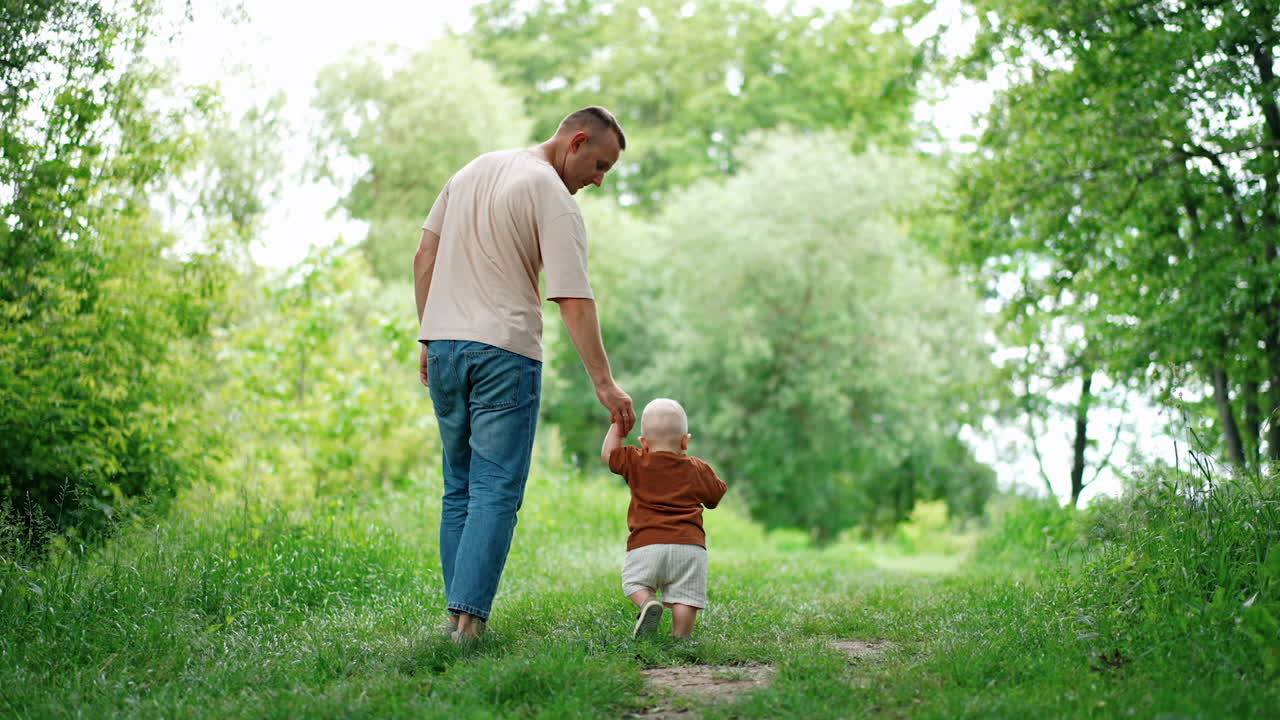 Dad leading his lovely little baby boy by the hand. Father and toddler son on the walk in summer.