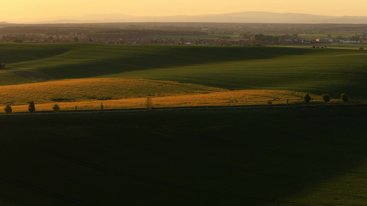 Aerial drone view of golden and green farmland at dusk with a small distant village, treelines and mountain silhouette on the horizon, Moravia, Czech Republic.