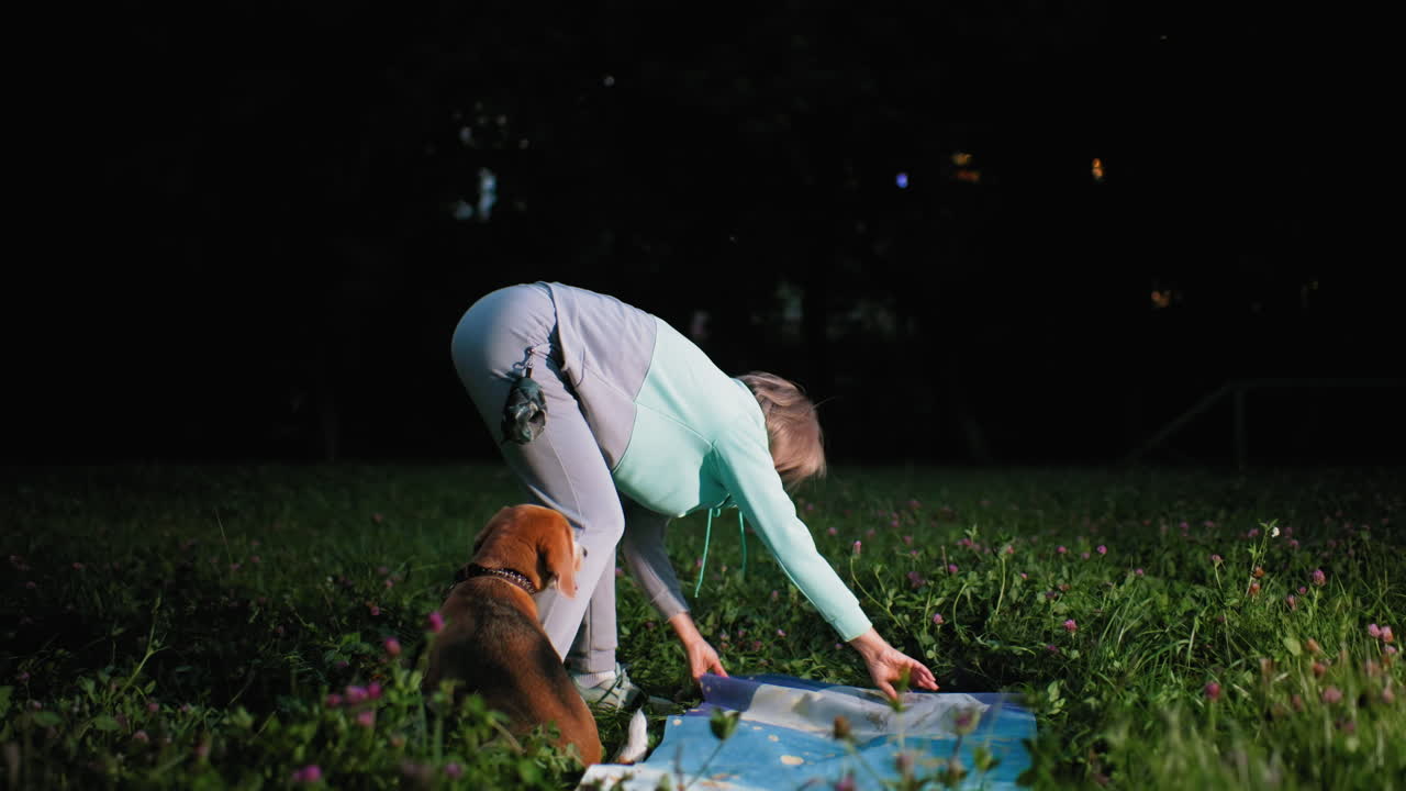 Dog owner spreads mat on green grass in evening park as small dog watches attentively creating calm peaceful outdoor moment filled with bonding, training, and preparation for relaxation