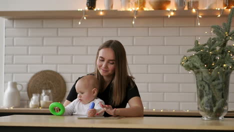 Mom-and-baby-sitting-at-a-table-in-a-white-bright-apartment.-Mom-presses-her-fingers-on-the-screen-of-the-tablet-computer,-and-the-son-of-a-baby-playing-with-colored-rings-from-the-pyramid