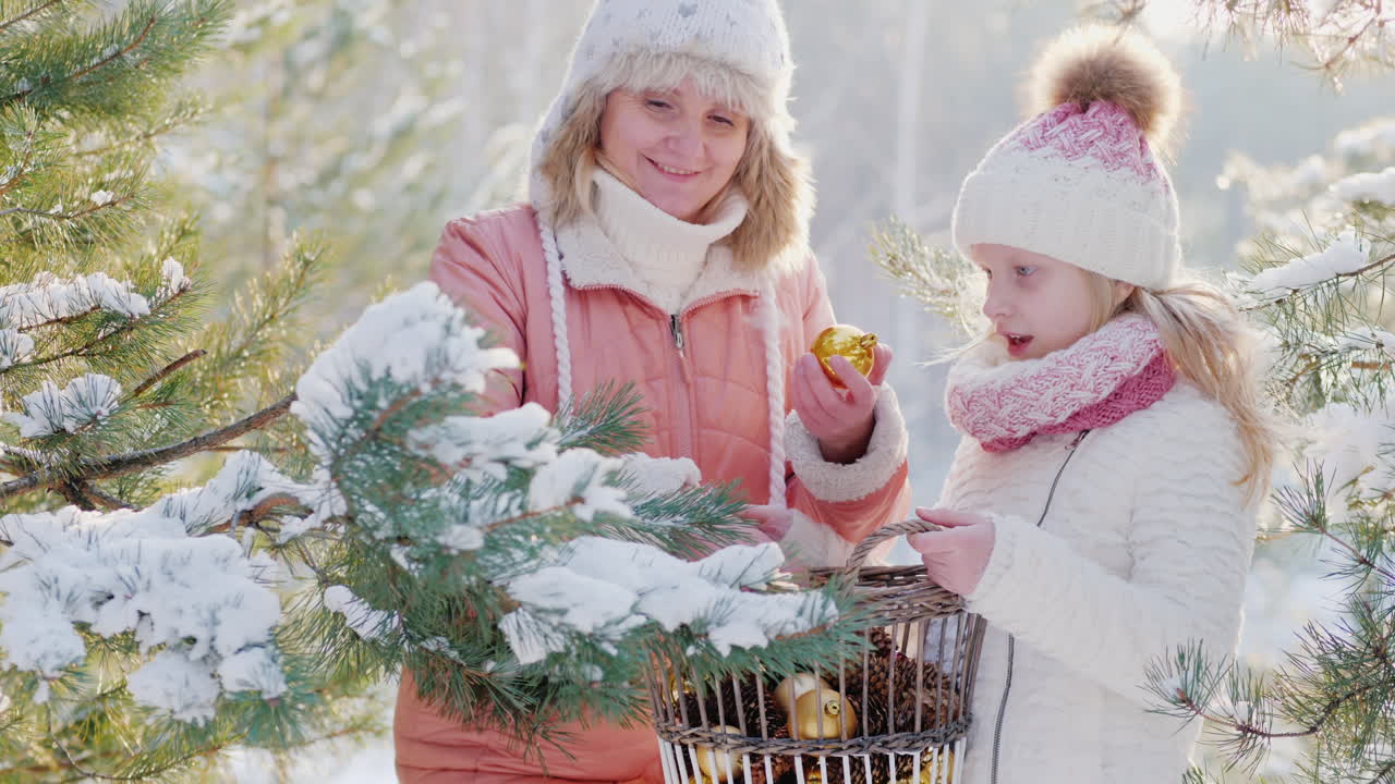 маленькая девочка и молодая мама украшают рождественскую елку декоративными шарами на заснеженном фоне