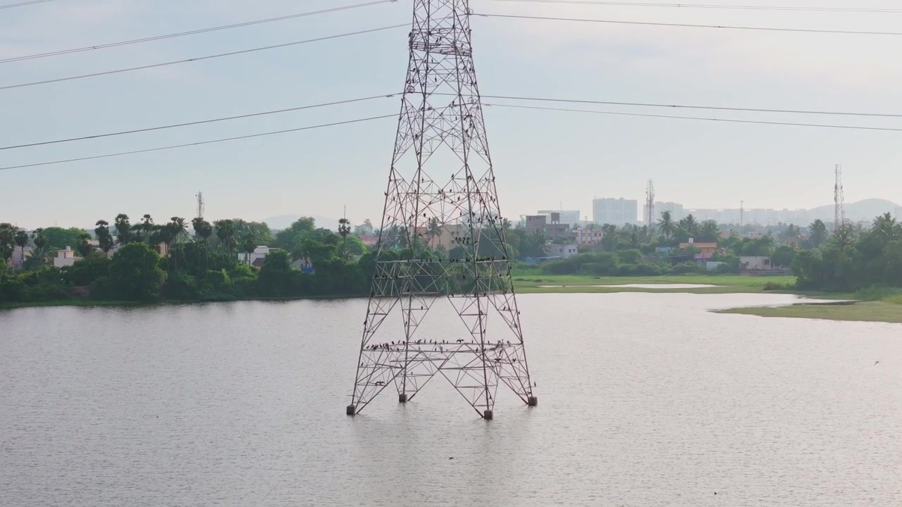 Aerial drone shot of power transmission tower standing in calm lake with overhead power lines, distant shoreline and urban skyline during daytime in Tamil Nadu, India.