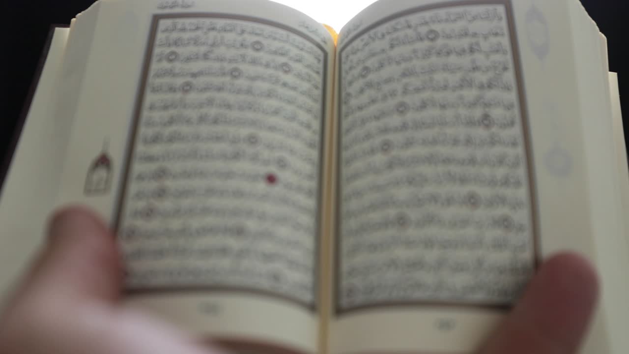 una persona levanta el corán y comienza a recitarlo, la toma se deja fuera de enfoque y se enfoca cuando el corán se acerca a la cámara
