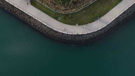 drone top down tracking follows families walking along ocean view promenade with grass patches