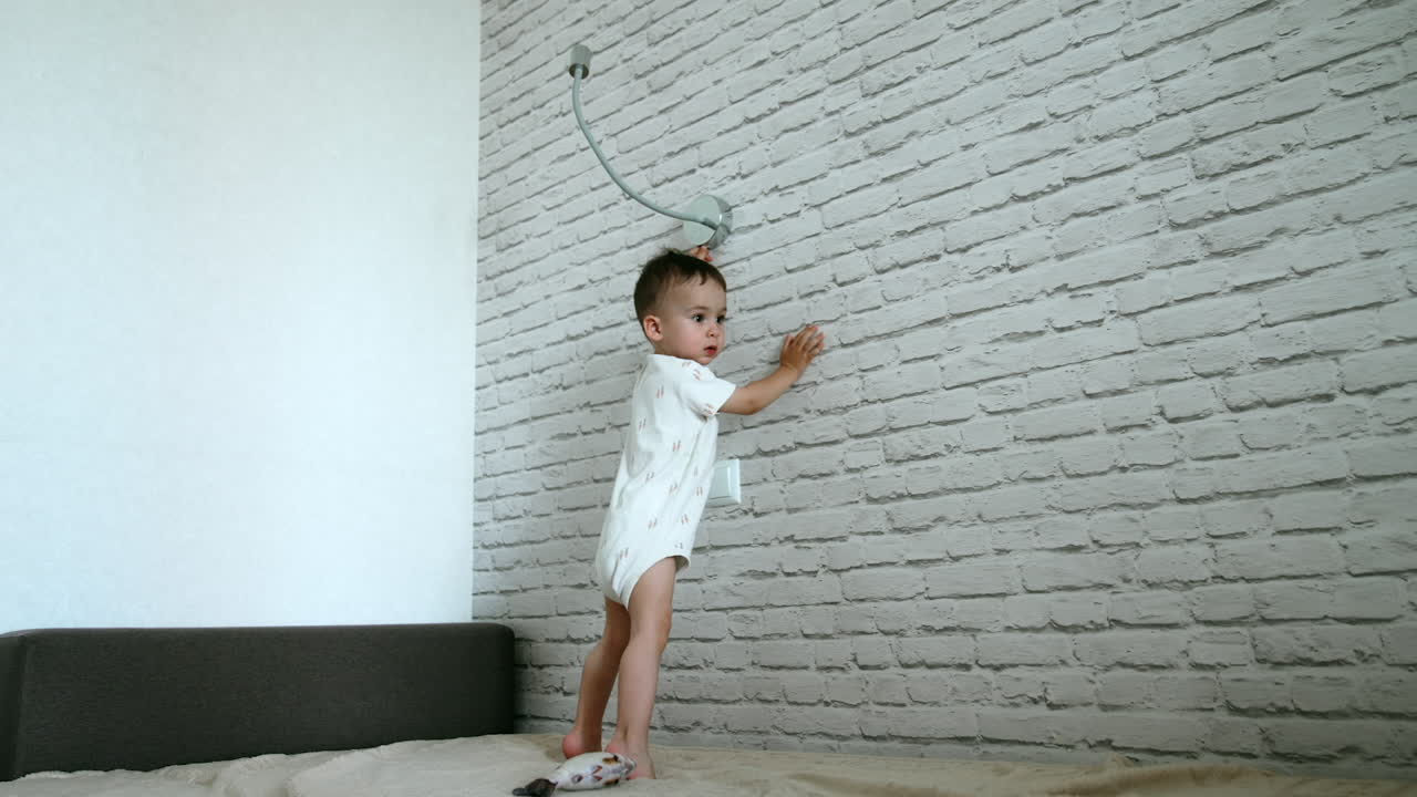 Pretty baby stands at the brick wall on the bed. Child wants to turn on the lamp but fails and jumps nervously. Low angle view.