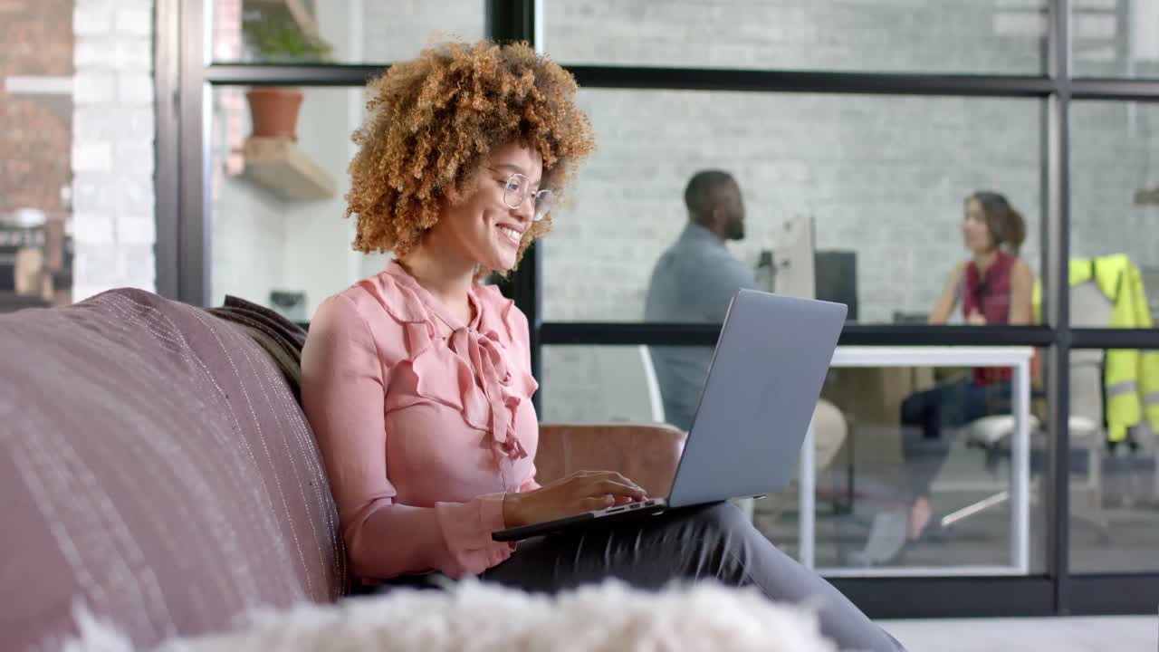 Happy biracial casual businesswoman using laptop on couch in office, copy space, slow motion