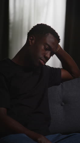 vertical video of unhappy and depressed young man sitting on sofa at home looking anxious and worried resting head on hand 5