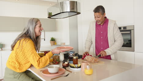 Preparing breakfast, multiracial senior couple in kitchen with fresh bread and orange juice, at home