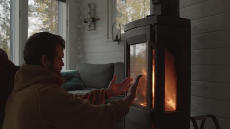 man sits in front of indoor fireplace warming his cold hands - medium close