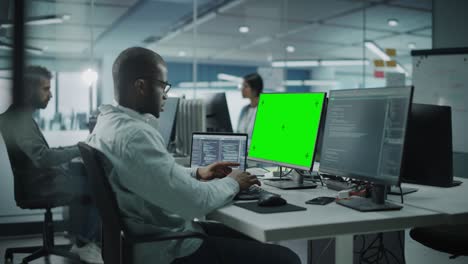 multi-ethnic office: black it programmer working on computer with green screen chroma key display. male software engineer developing app, program, video game. terminal with code language. static shot