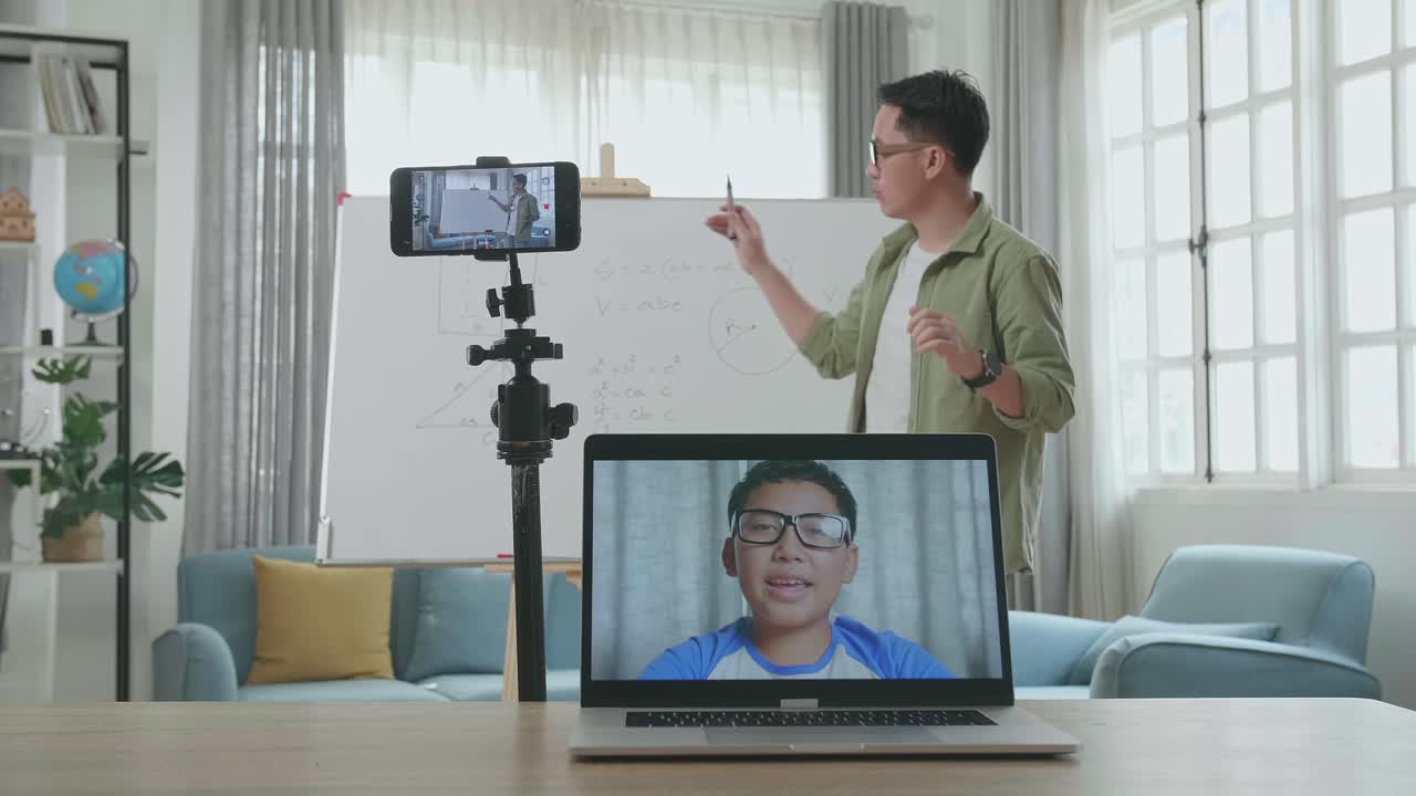 Asian Male Teacher With Glasses Shoots Video By Smartphone Camera And Having Video Call On Laptop While Teaching Math At Home