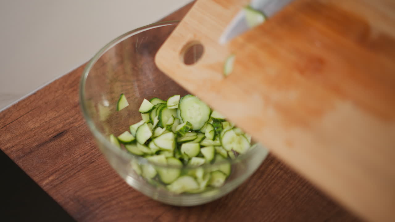 Close up of person pouring freshly sliced cucumber into transparent glass bowl on wooden kitchen counter, with cutting board and knife visible