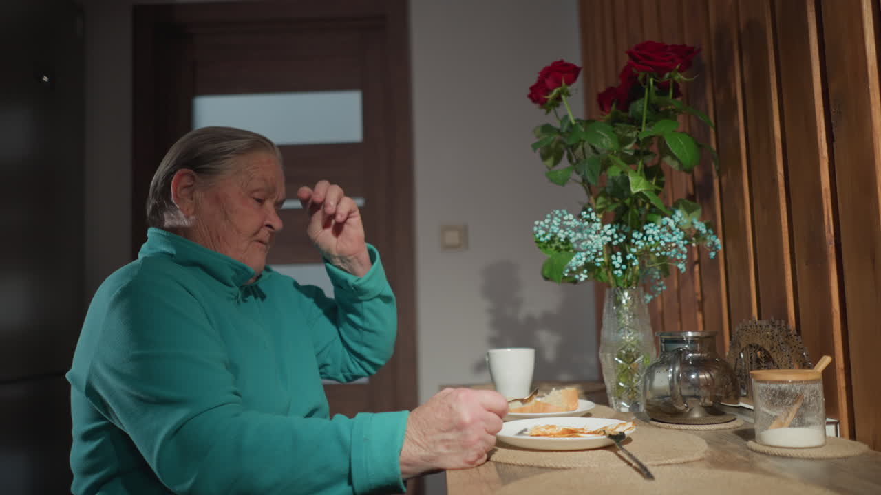 elderly woman wearing a green fleece cloth is enjoying breakfast by sitting at a table. She is taking a break from eating bread and eggs, while a beautiful flower vase decorates her table