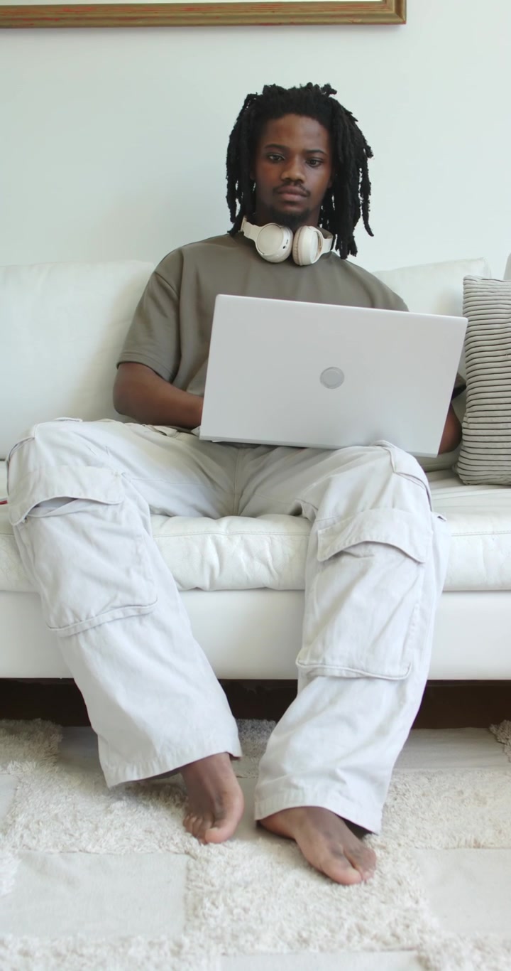 Vertical video: African American man starting on white sofa with laptop open and headset, working