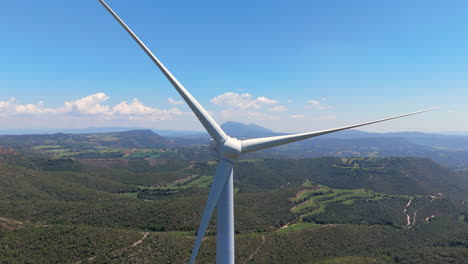 cinematic drone shot retreating from a large wind turbine spinning over green mountains, revealing panoramic views of the valley and distant peaks under clear skies