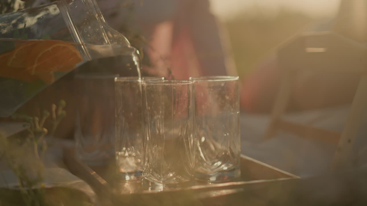 Close-up of someone pouring infused water from pitcher into three glasses under warm sunlight, orange slices and mint leaves visible in water, with blur background