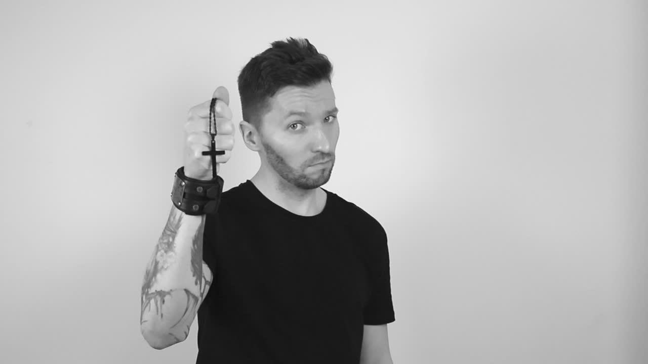 Grayscale footage of a tattooed man lifting up a holy black cross on a chain while it moves and dangles in his hand in slow motion. The man nods and shows that he approves faith and religion.