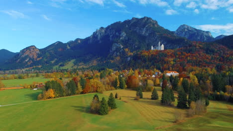 Toma-En-ángulo-Curvo-De-Drones-Deslizándose-Y-Volando-Sobre-Casas-De-Pueblo-Hacia-El-Castillo-En-La-Colina-Sobre-El-Pintoresco-Campo-De-Otoño-En-La-Tarde-Cerca-Del-Castillo-De-Neuschwanstein-En-Alemania,-Europa,-Vista-Amplia