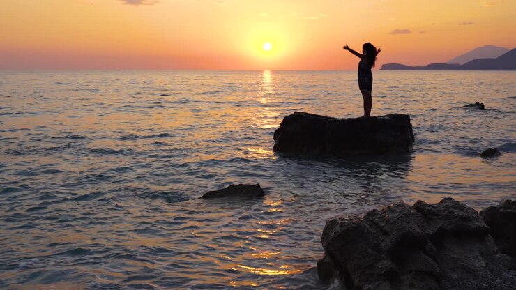 Exercício de yoga realizado por uma jovem mulher em pé sobre uma rocha banhada pelo mar, com um céu laranja ao pôr do sol como fundo.
