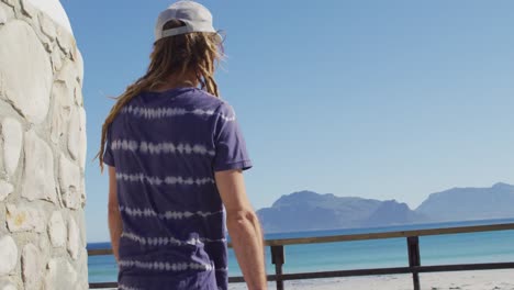 video of thoughtful caucasian man with dreadlocks standing on sunny promenade looking out to sea