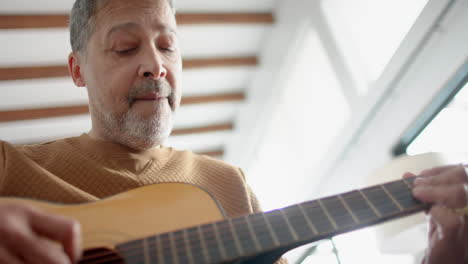 low angle of focused senior biracial man sitting playing guitar at home, slow motion